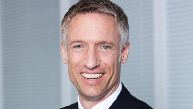 Ein lächelnder Mann im Anzug vor einem hellen Hintergrund.