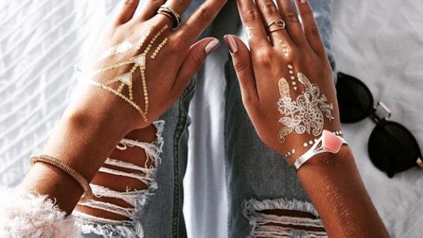 Die Hände einer Person mit goldenen und silbernen Tattoos liegen auf zerrissenen Jeans.