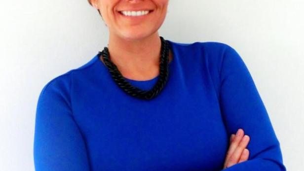 Eine Frau mit roten Brillengläsern und blauem Oberteil lächelt mit verschränkten Armen.