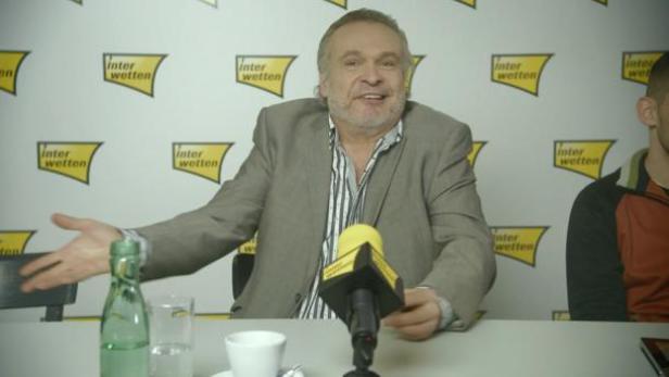 Ein Mann in einem grauen Anzug sitzt an einem Tisch mit einem Mikrofon vor einer Wand mit „Interwetten“-Logos.