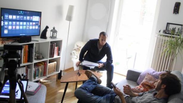 Zwei Männer in einem Wohnzimmer, einer sitzt auf dem Sofa und benutzt ein Handy, der andere sitzt auf einem Stuhl mit Papieren in der Hand.