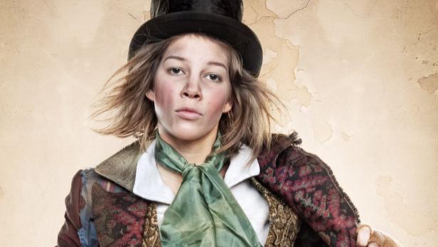 Eine junge Person in historischer Kleidung und Zylinder vor einem beigen Hintergrund.