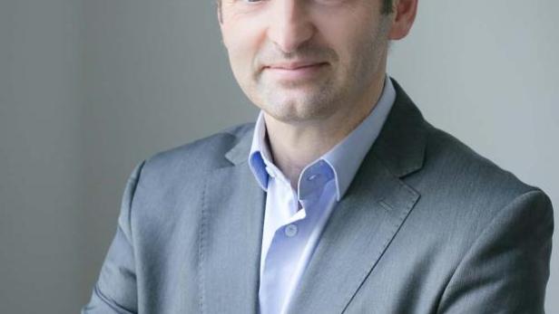 Ein Mann im grauen Anzug steht mit verschränkten Armen vor einem grauen Hintergrund.