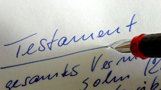 Eine Feder schreibt das Wort „Testament“ auf ein Blatt Papier.