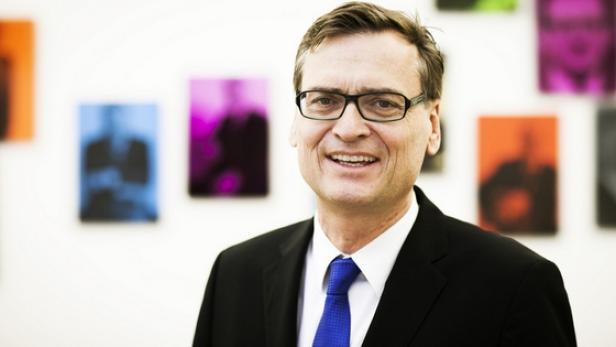 Ein lächelnder Mann mit Brille, Anzug und Krawatte vor farbigen Bildern.
