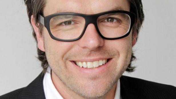 Ein lächelnder Mann mit Brille und Anzug vor einem neutralen Hintergrund.