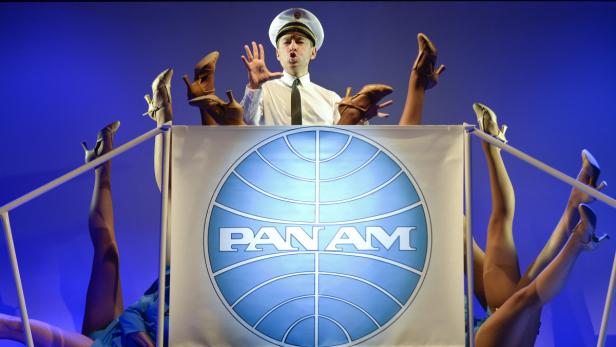 Ein Mann in Pilotenuniform steht hinter einem Pan-Am-Banner, umgeben von hochgestreckten Beinen in High Heels.
