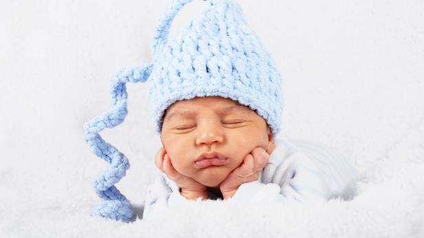 Ein schlafendes Baby mit blauer Zipfelmütze liegt auf einer weißen Decke.