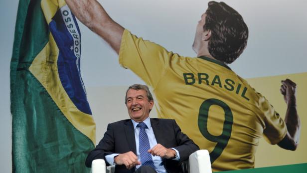 Ein lachender Mann im Anzug sitzt vor einer brasilianischen Flagge und einem Bild eines Fußballspielers.