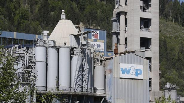 Eine Zementfabrik mit Silos und einem hohen Turm vor einem bewaldeten Hügel.