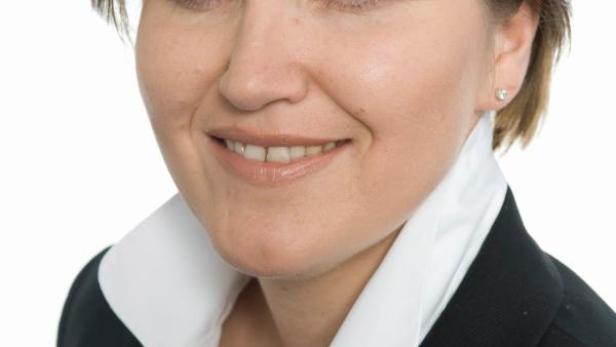 Porträt einer lächelnden Frau mit kurzem, blonden Haar und blauen Augen, die einen Blazer trägt.