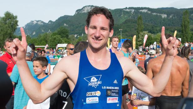 Ein lächelnder Athlet in Sportkleidung zeigt mit beiden Händen nach oben.