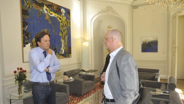 Zwei Männer unterhalten sich in einem eleganten Raum mit Kunstwerken an den Wänden.