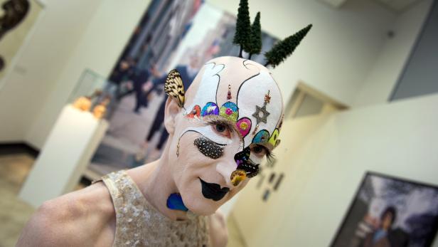Eine Person mit aufwendigem Make-up und kleinen Bäumen auf dem Kopf in einer Galerie.