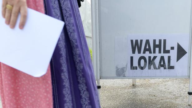 Eine Frau in Tracht hält einen Stimmzettel vor einem Schild, das auf ein Wahllokal hinweist.