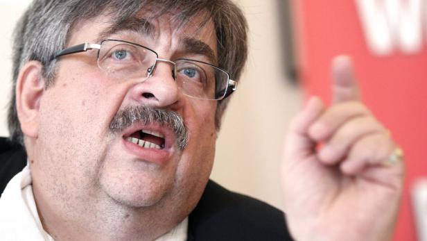 Ein Mann mit Brille und Schnurrbart gestikuliert mit erhobenem Zeigefinger.