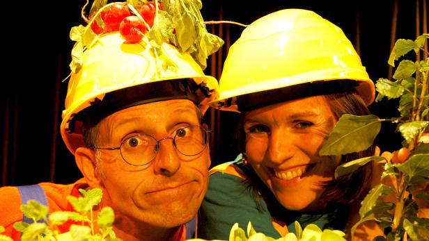 Zwei Personen mit Bauhelmen, einer mit Radieschen geschmückt, lächeln in die Kamera.