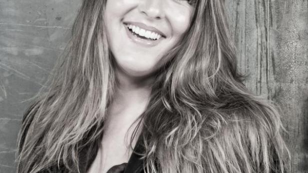 Eine lächelnde Frau mit langen Haaren vor einem grauen Hintergrund.