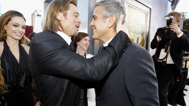 Brad Pitt und George Clooney umarmen sich auf dem roten Teppich, während Angelina Jolie lächelt.