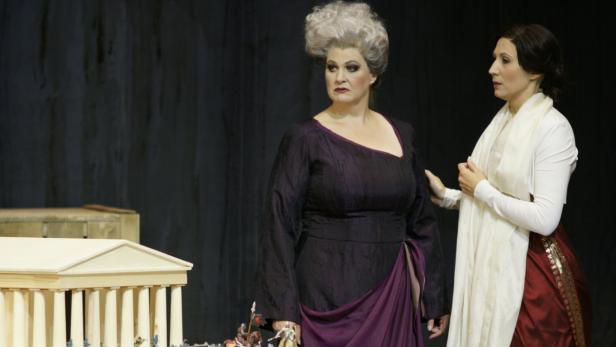 Zwei Frauen in Kostümen stehen auf einer Bühne vor einer Miniaturkulisse eines griechischen Tempels.