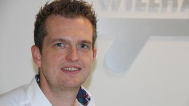 Ein Mann mit kurzem braunem Haar und einem weißen Hemd lächelt in die Kamera.