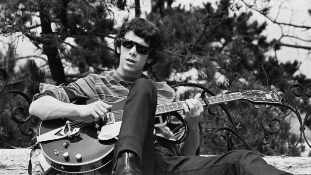 Lou Reed sitzt mit Sonnenbrille und Gitarre im Freien.