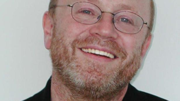 Ein lächelnder Mann mit Brille und Bart vor einem weißen Hintergrund.