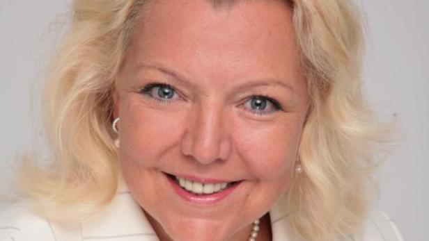 Porträt einer lächelnden Frau mit blonden Haaren, Perlenkette und weißem Blazer.