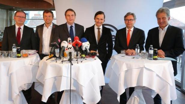 Sechs Männer stehen hinter Tischen mit Mikrofonen bei einer Pressekonferenz.