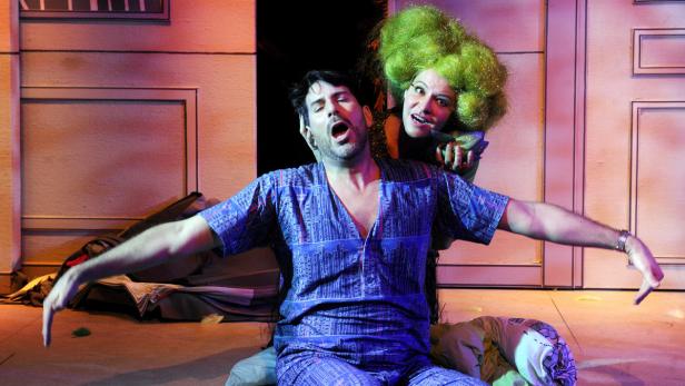 Ein Mann im Schlafanzug wird von einer Frau mit grüner Perücke überrascht.