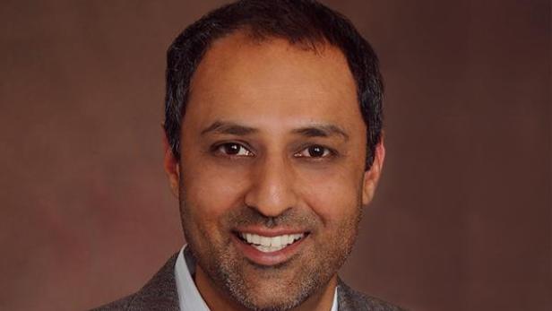 Ein lächelnder Mann mit dunklen Haaren und Bart vor einem braunen Hintergrund.