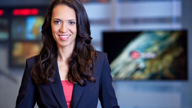 Eine lächelnde Frau in einem Blazer steht vor Bildschirmen in einem Studio.