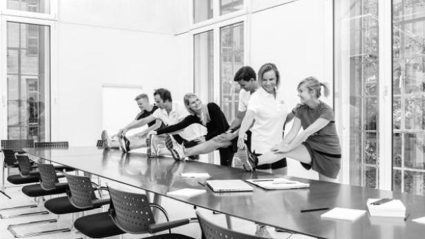 Eine Gruppe von Menschen dehnt sich an einem langen Konferenztisch in einem hellen Raum.