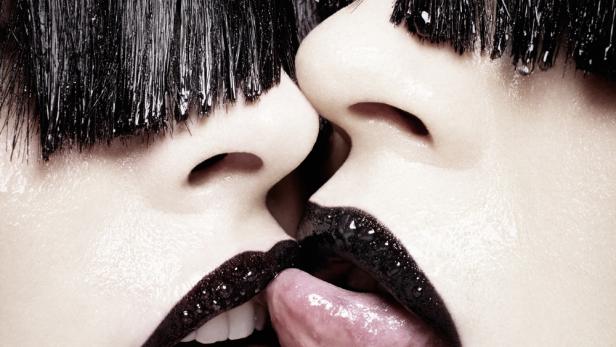 Zwei Gesichter mit schwarzem Lippenstift und ausgestreckten Zungen berühren sich.