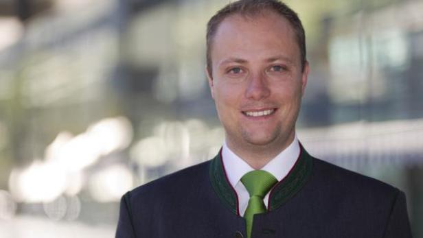 Ein lächelnder Mann im Trachtenanzug mit grünem Schlips vor einem modernen Gebäude.