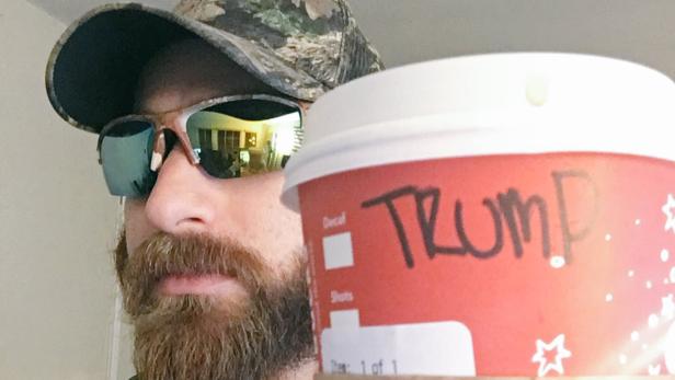 Ein Mann mit Bart und Kappe hält einen Kaffeebecher mit der Aufschrift „Trump“.