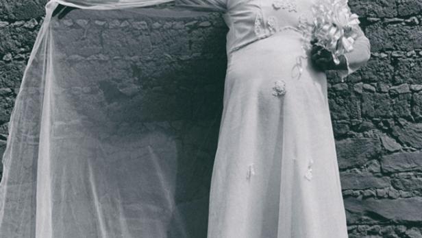 Eine Braut mit Totenkopfmaske und Brautstrauß steht vor einer Mauer.