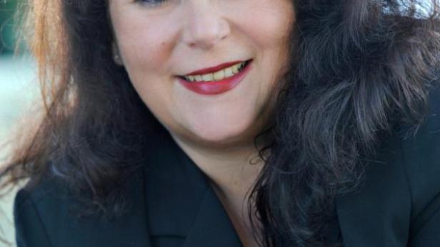 Eine Frau mit dunklen, lockigen Haaren und einem schwarzen Blazer lächelt in die Kamera.