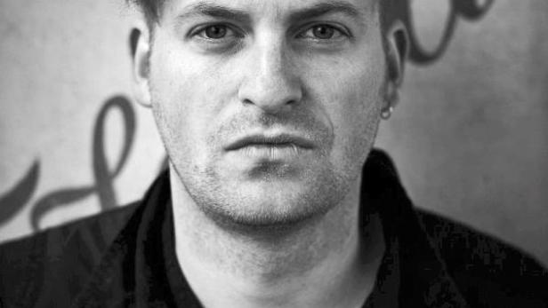 Schwarzweiß-Porträt eines Mannes mit kurzer Frisur und Jacke vor einer Wand mit Schriftzug.