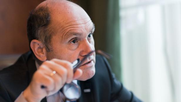 Ein Mann im Anzug hält eine Brille in der Hand und schaut nachdenklich zur Seite.