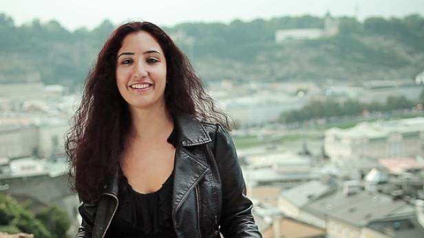Eine lächelnde Frau mit lockigem Haar und Lederjacke vor einer Stadtansicht.