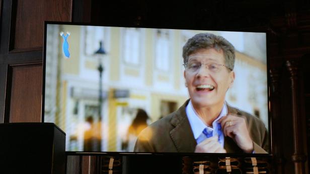 Ein Mann mit Brille und braunem Sakko bindet seine Krawatte vor einer Stadtkulisse.