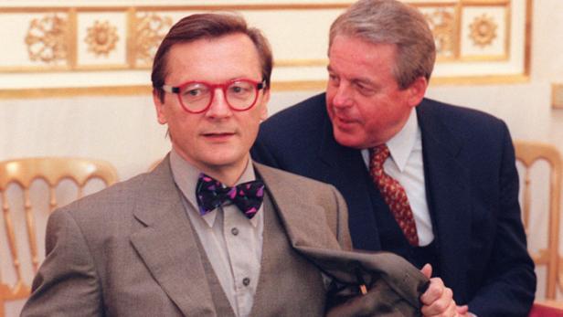 Ein Mann mit roter Brille und Fliege, neben ihm ein Mann im Anzug.