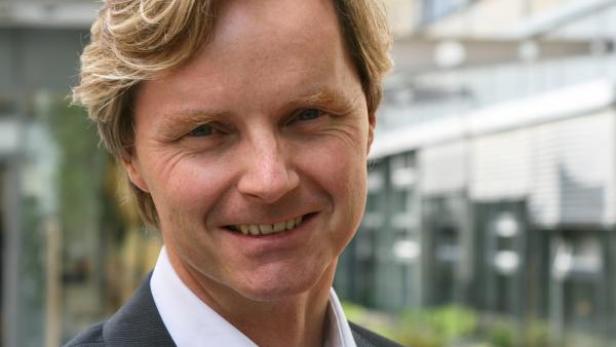 Ein Mann mit blonden Haaren und einem dunklen Anzug lächelt in die Kamera.