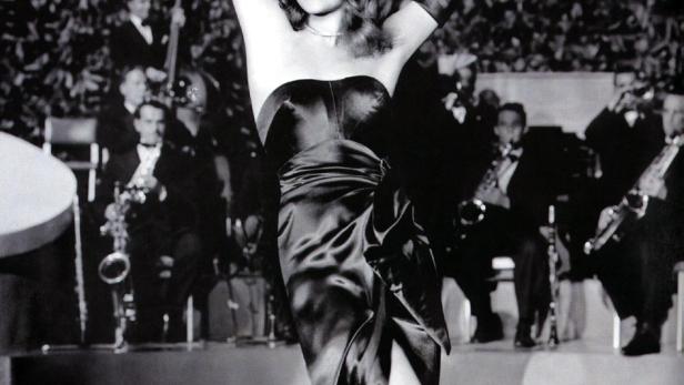 Rita Hayworth in einem schwarzen Kleid mit hoch erhobenen Armen, begleitet von einer Band.