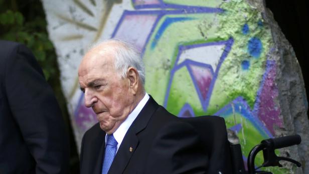 Helmut Kohl sitzt in einem Rollstuhl vor einem bemalten Mauerstück.