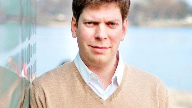 Ein Mann mit braunen Haaren und einem beigen Pullover posiert vor einem Gewässer.