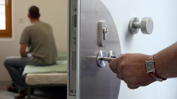 Ein Mann schließt eine schwere Tür auf, während im Hintergrund eine Person auf einem Bett sitzt.