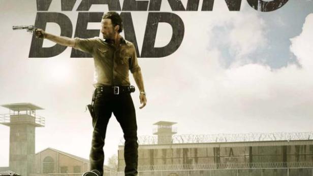 Ein Mann steht mit einer Waffe auf einem Fahrzeug vor einem Gefängnis, im Hintergrund der Schriftzug „The Walking Dead“.
