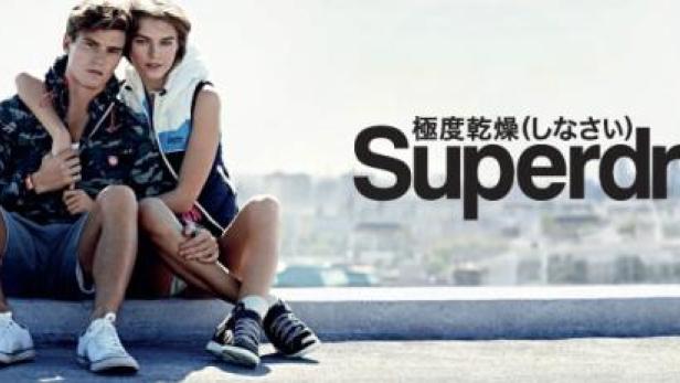 Ein junges Paar sitzt vor einer Stadtkulisse, im Hintergrund das Logo von Superdry.